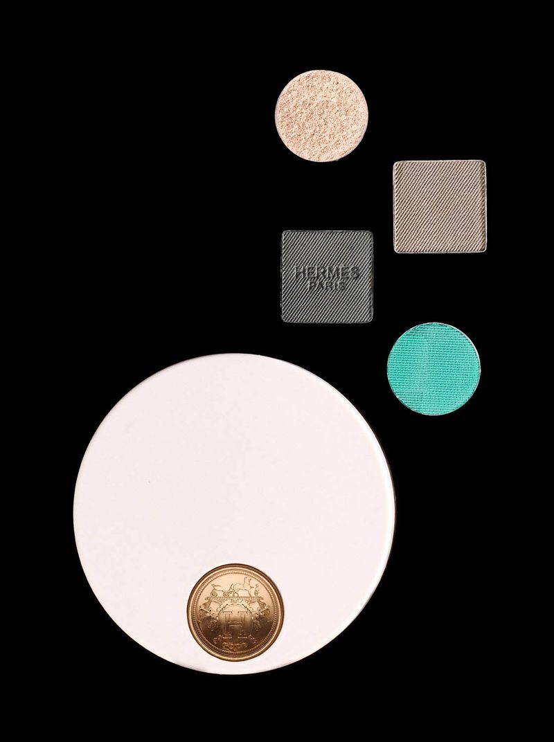 Hermès Paris makeup compact and four colorful eyeshadow pans arranged on a black background in a beauty still life shot.
