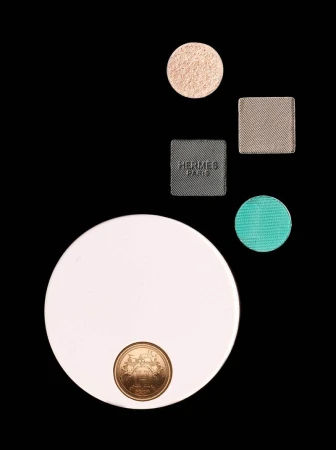 Hermès Paris makeup compact and four colorful eyeshadow pans arranged on a black background in a beauty still life shot.