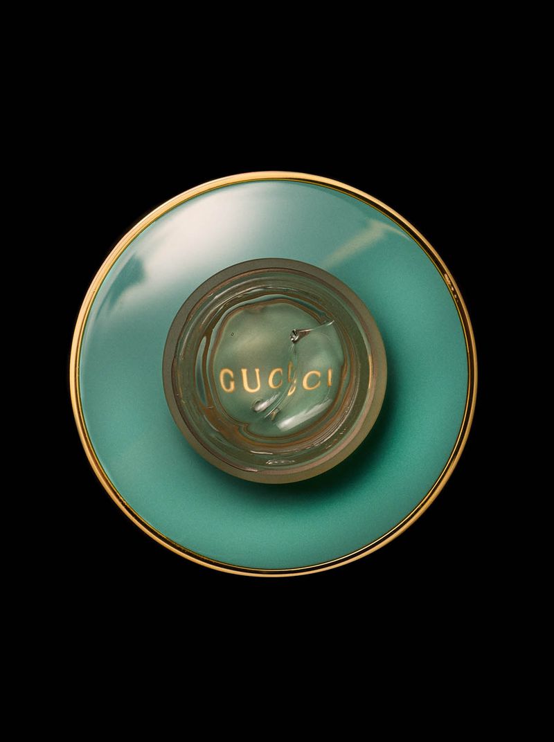 Overhead shot of a Gucci cosmetic jar with a teal base and gold trim against a black background, highlighting the container's design.