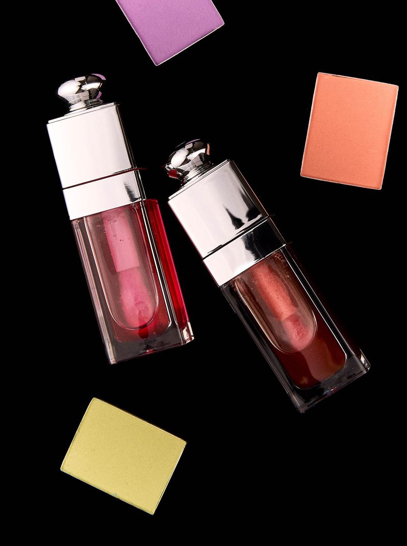 Still life of two lip glosses in clear, rectangular bottles with silver caps, surrounded by cosmetic eyeshadow squares on black backdrop.