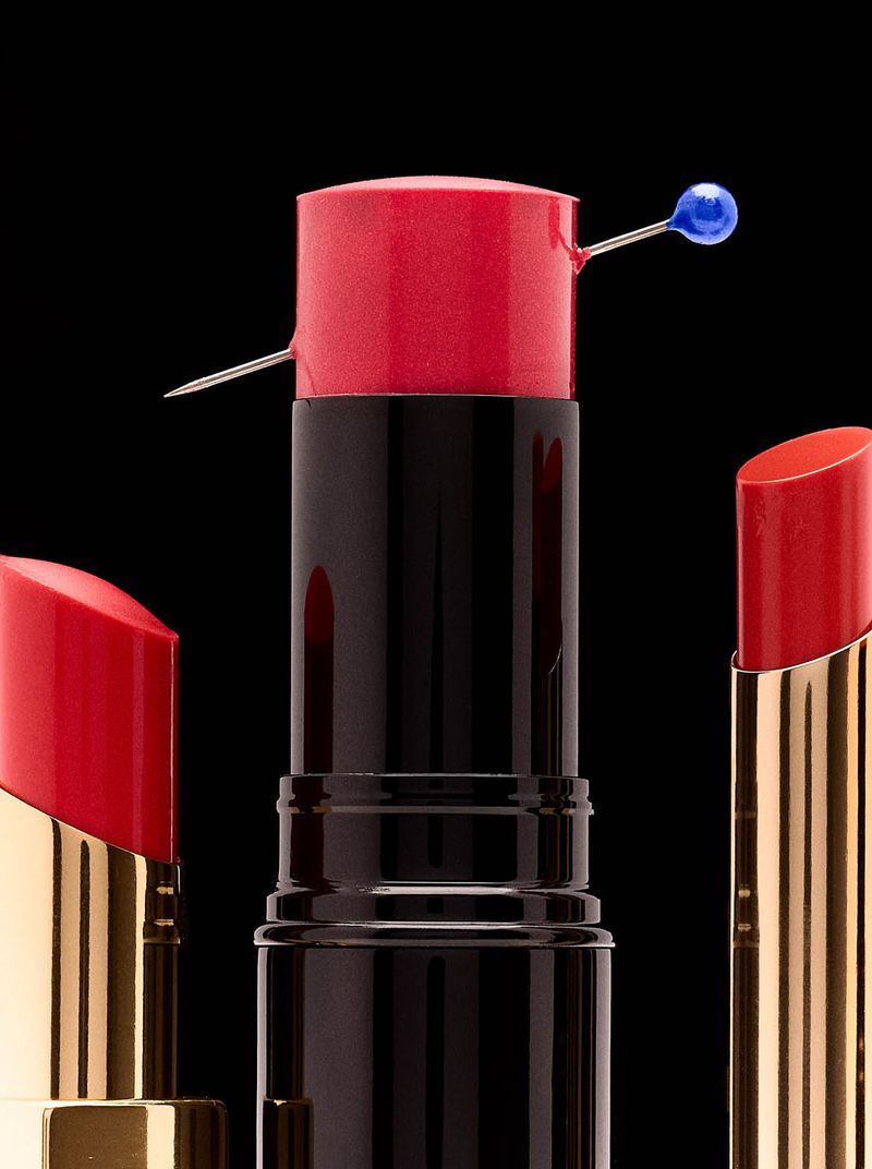 Close-up studio shot of red lipsticks in gold and black tubes against a black backdrop, one featuring a sewing pin through the lipstick.