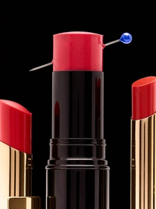 Close-up product photography of three red lipsticks arranged against a stark black background with a blue pin piercing the central tube