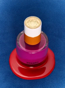 Close-up overhead shot of a colourful Hermes Beauty lipstick bottle resting on a blue textured surface.