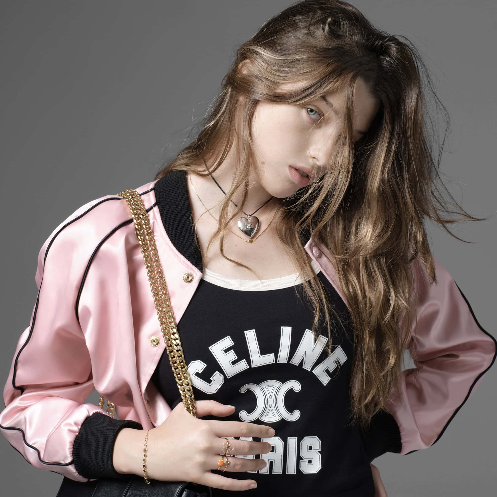 Fashion portrait of a young woman with long wavy hair wearing a pink jacket, black shirt, and heart necklace against a grey background.