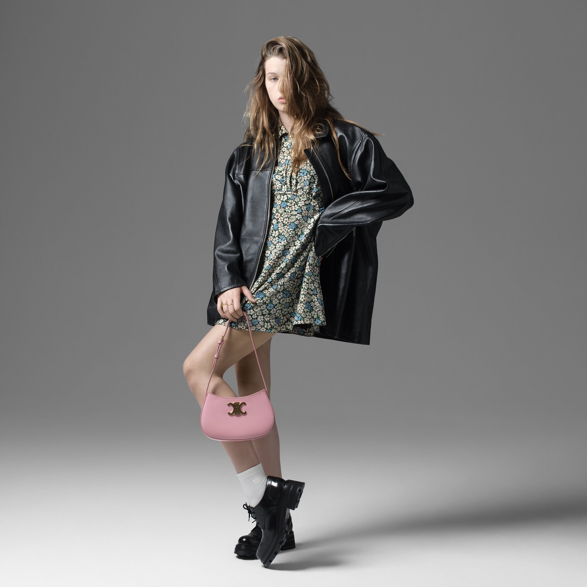 A young woman in a floral dress and oversized leather jacket poses in a studio with a pink purse and black boots, full shot.