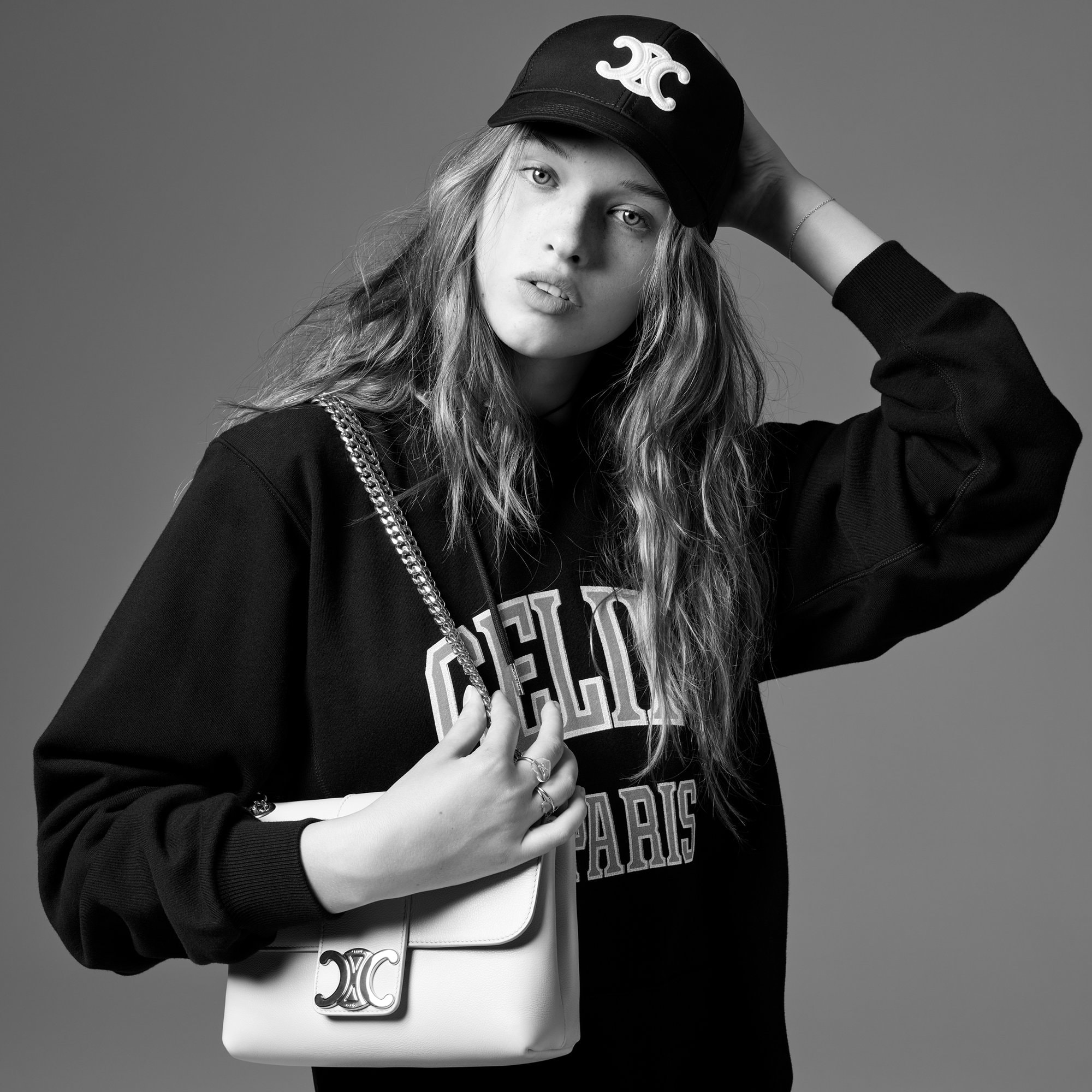 A young adult female poses in a black sweatshirt and ball cap, accessorized with a white purse with a chain strap, against a grey backdrop.