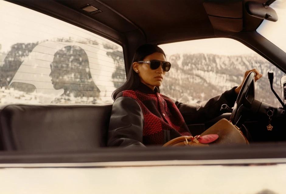 A woman in sunglasses sits in the driver's seat of a car, with snow-covered mountains visible through the windshield, casting natural light.