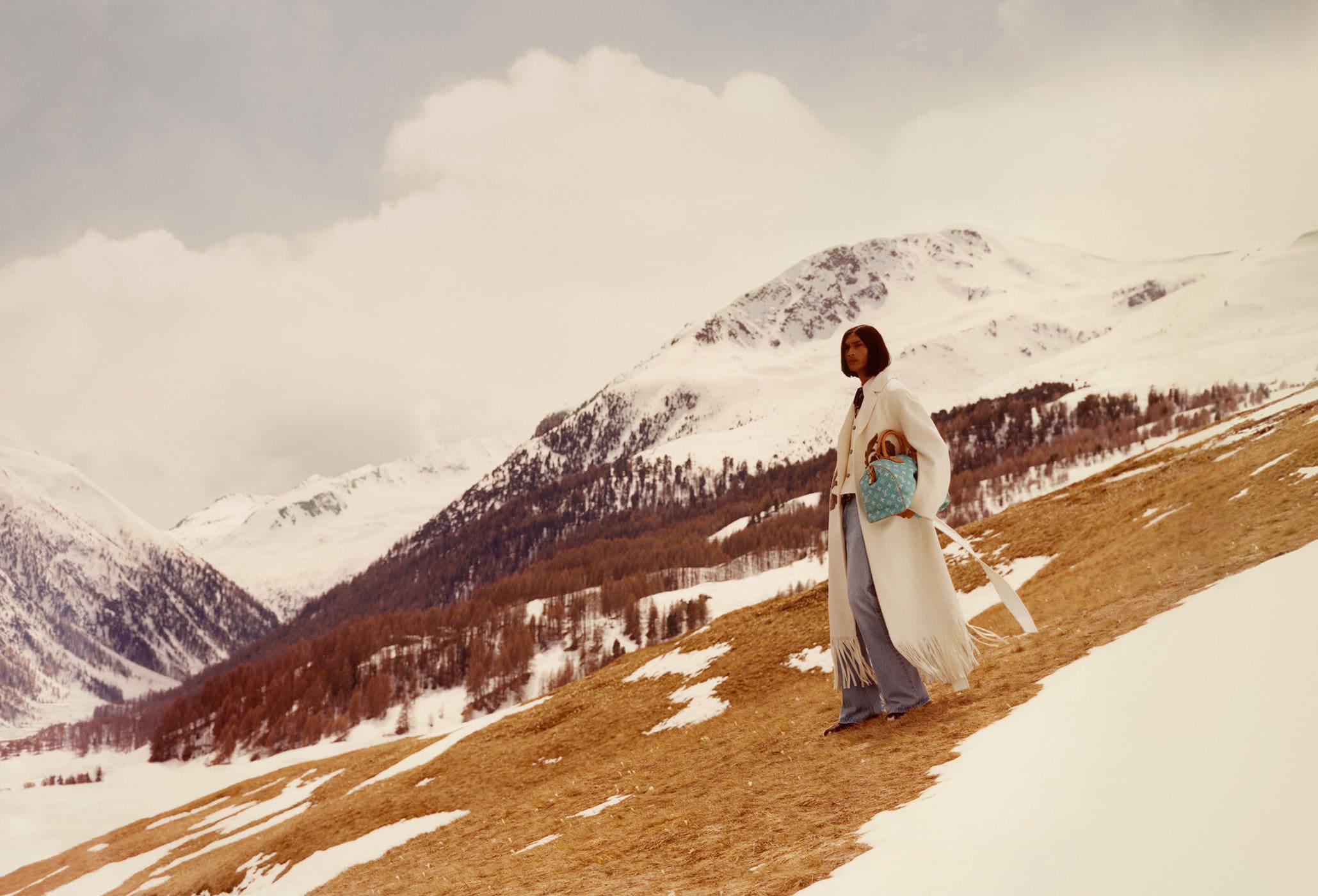 A woman in a white coat and jeans stands on a hillside with snow-covered mountains in the background, carrying a blue patterned handbag.