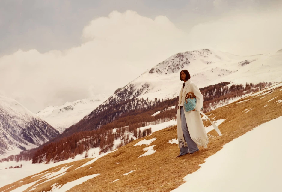 A woman in a white coat and jeans stands on a hillside with snow-covered mountains in the background, carrying a blue patterned handbag.