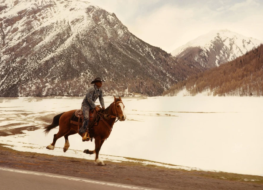 A man in a cowboy hat rides a horse at a gallop on a road with a snowy mountain range in the background on a cloudy day.
