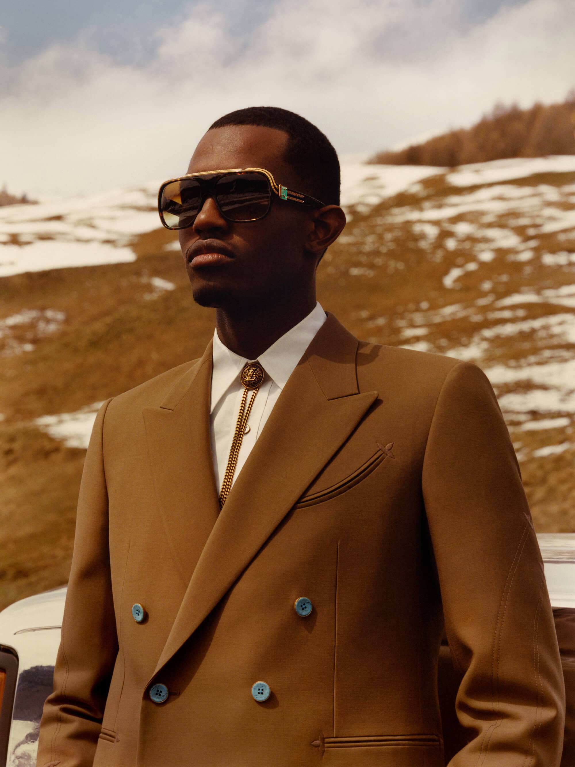 A stylish African American man in a brown suit and sunglasses poses confidently against a backdrop of snow-dusted hills.