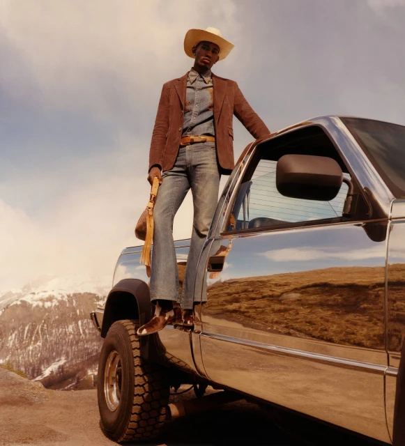 Dark-skinned male model in cowboy hat and boots stands on a silver truck in a mountainous landscape under a cloudy sky.