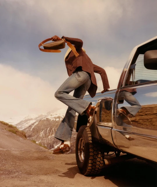 Fashion shot of a stylish adult wearing a brown jacket and jeans climbing on the shiny hood of a silver SUV near mountains and sky.