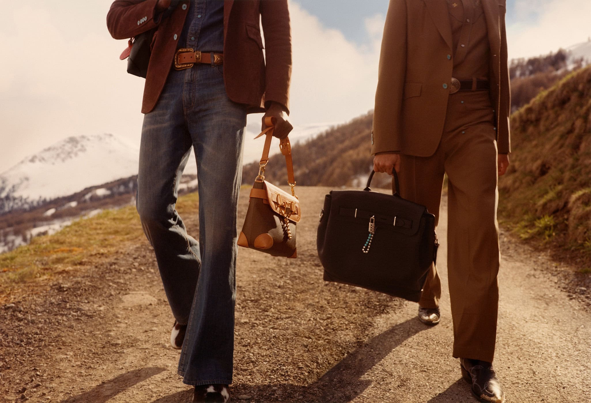 Two stylish men walk on a rural path, carrying a handbag and briefcase, with snow-capped mountains visible in the background.