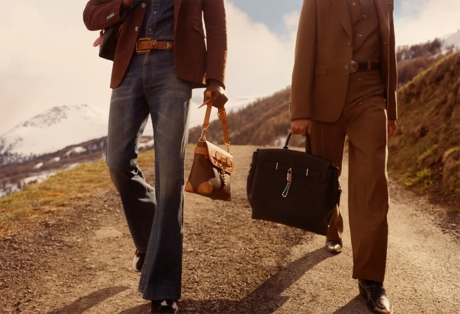 Two stylish men walk on a rural path, carrying a handbag and briefcase, with snow-capped mountains visible in the background.