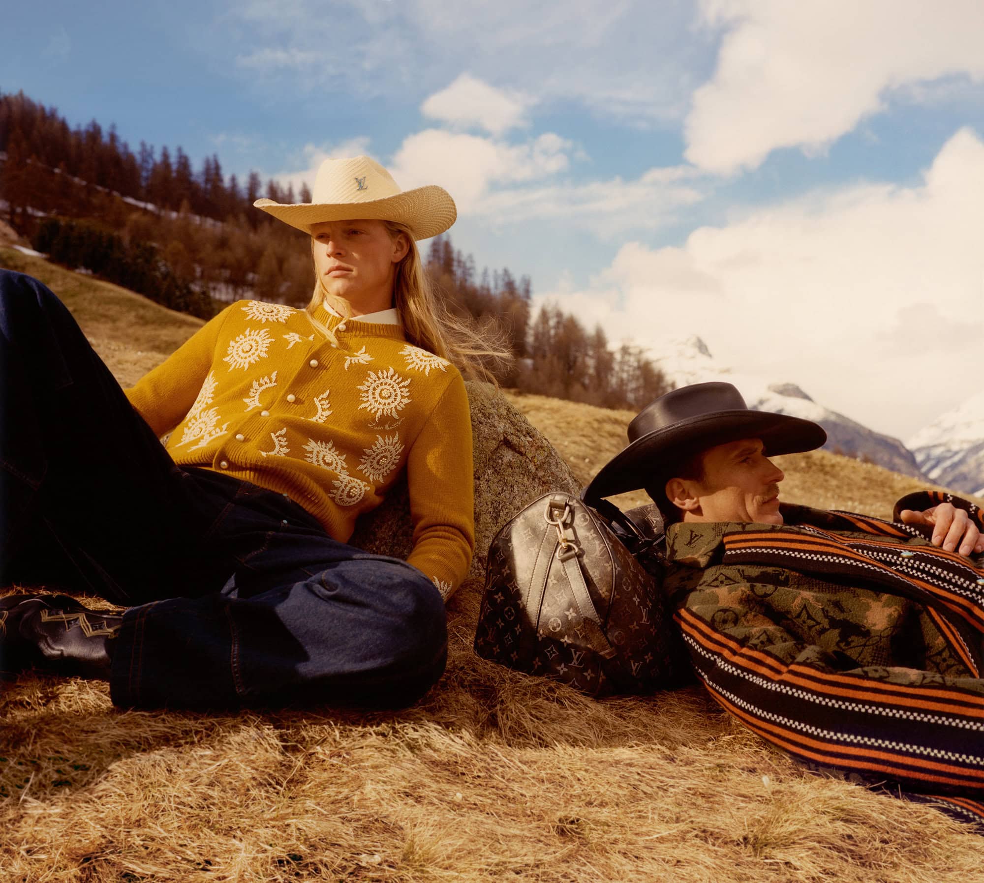 Two adult male models pose outdoors wearing western-style clothing and cowboy hats with a Louis Vuitton travel bag in a field setting.