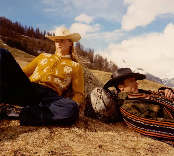 Two adult male models pose outdoors wearing western-style clothing and cowboy hats with a Louis Vuitton travel bag in a field setting.