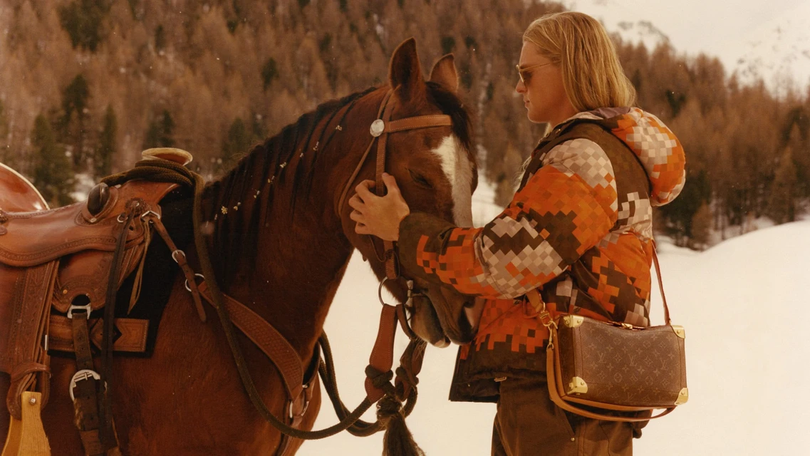 Person with long blond hair petting a brown horse wearing a saddle in a snowy landscape with trees. Model wearing jacket and Louis Vuitton bag.
