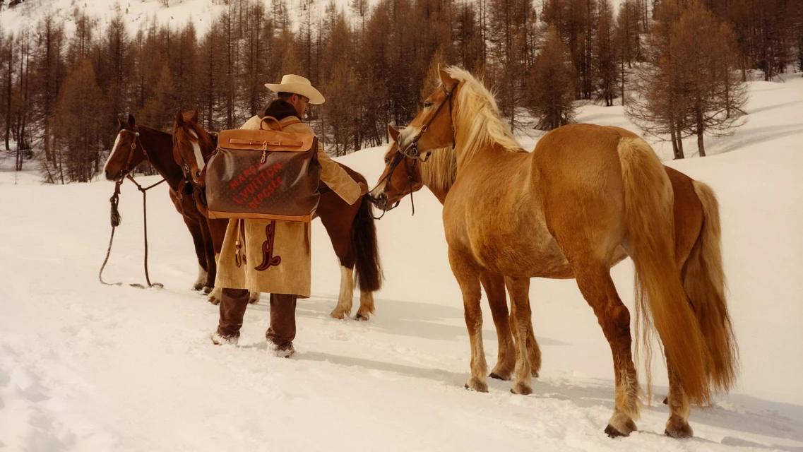A man in cowboy attire with a Louis Vuitton backpack stands in the snowy mountains with several brown horses in an outdoor, daytime setting.