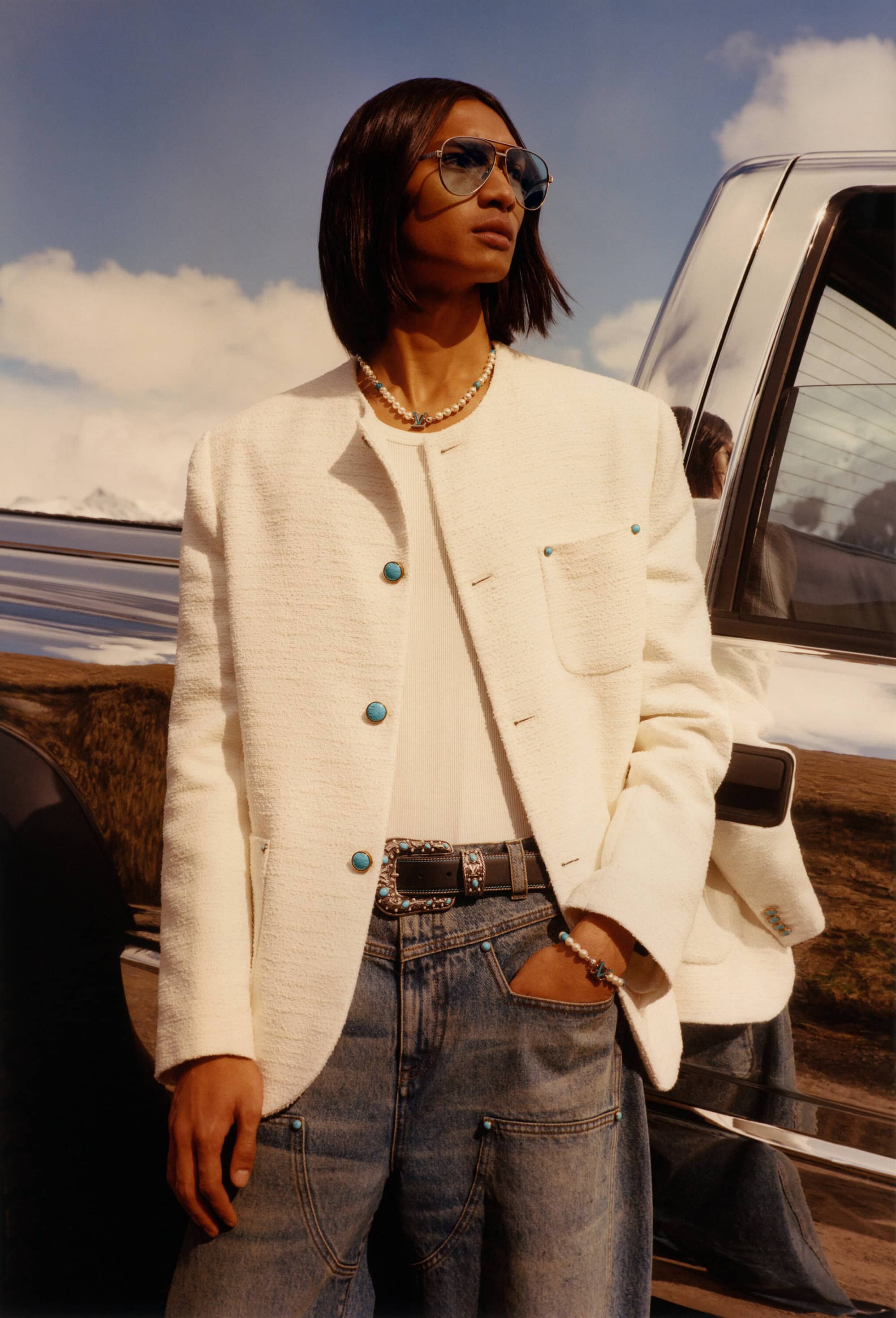 A stylish adult male poses in a white jacket and blue jeans near a truck against a blue sky with a reflective surface and a sunny day.