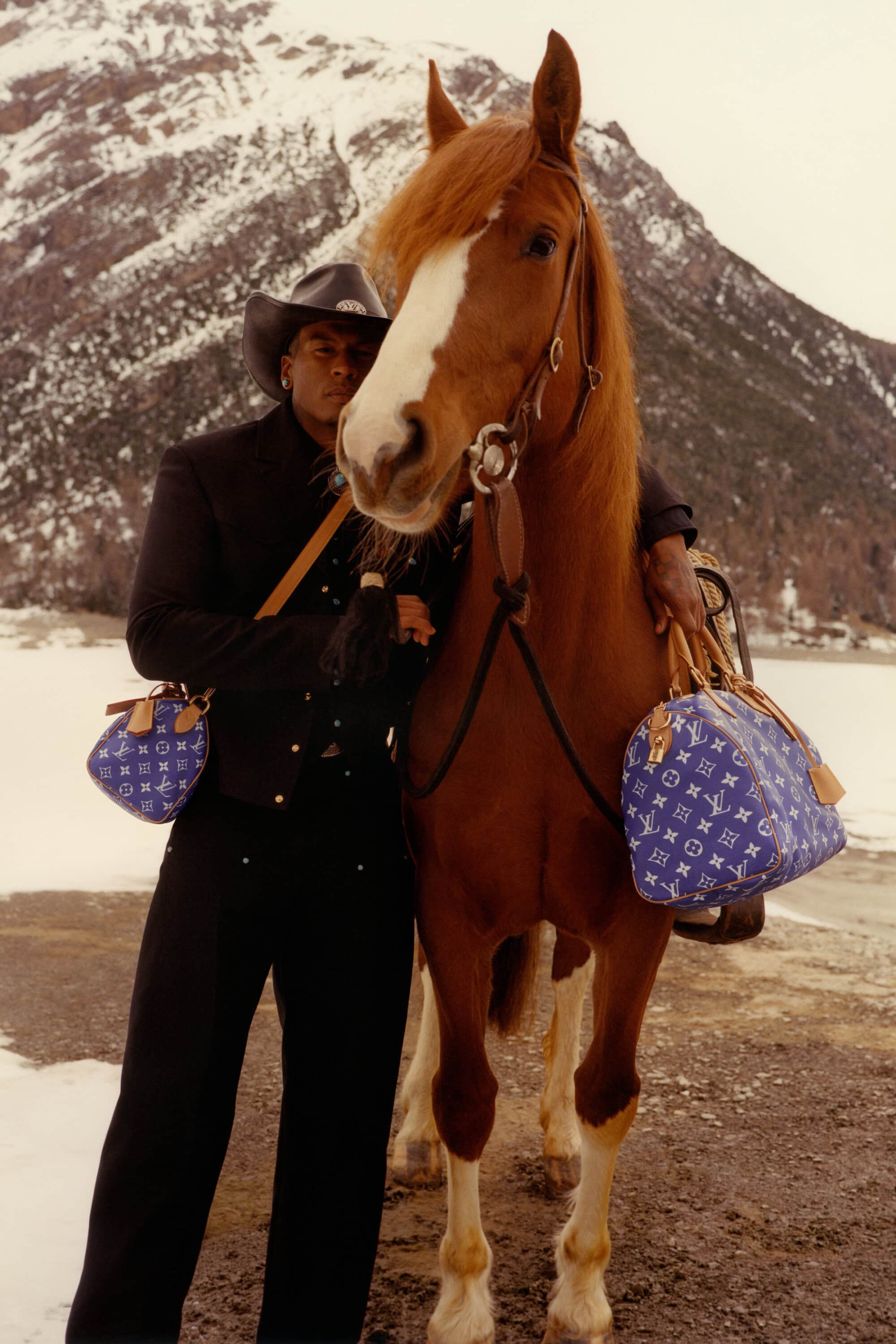 African-descent man in cowboy hat poses with brown horse in snowy mountains, holding designer bags. Travel, fashion, and outdoor lifestyle.