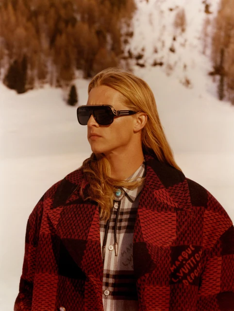 Man with sunglasses wearing a red and black patterned coat posing in a snowy mountain landscape on a sunny day, fashion editorial shot.