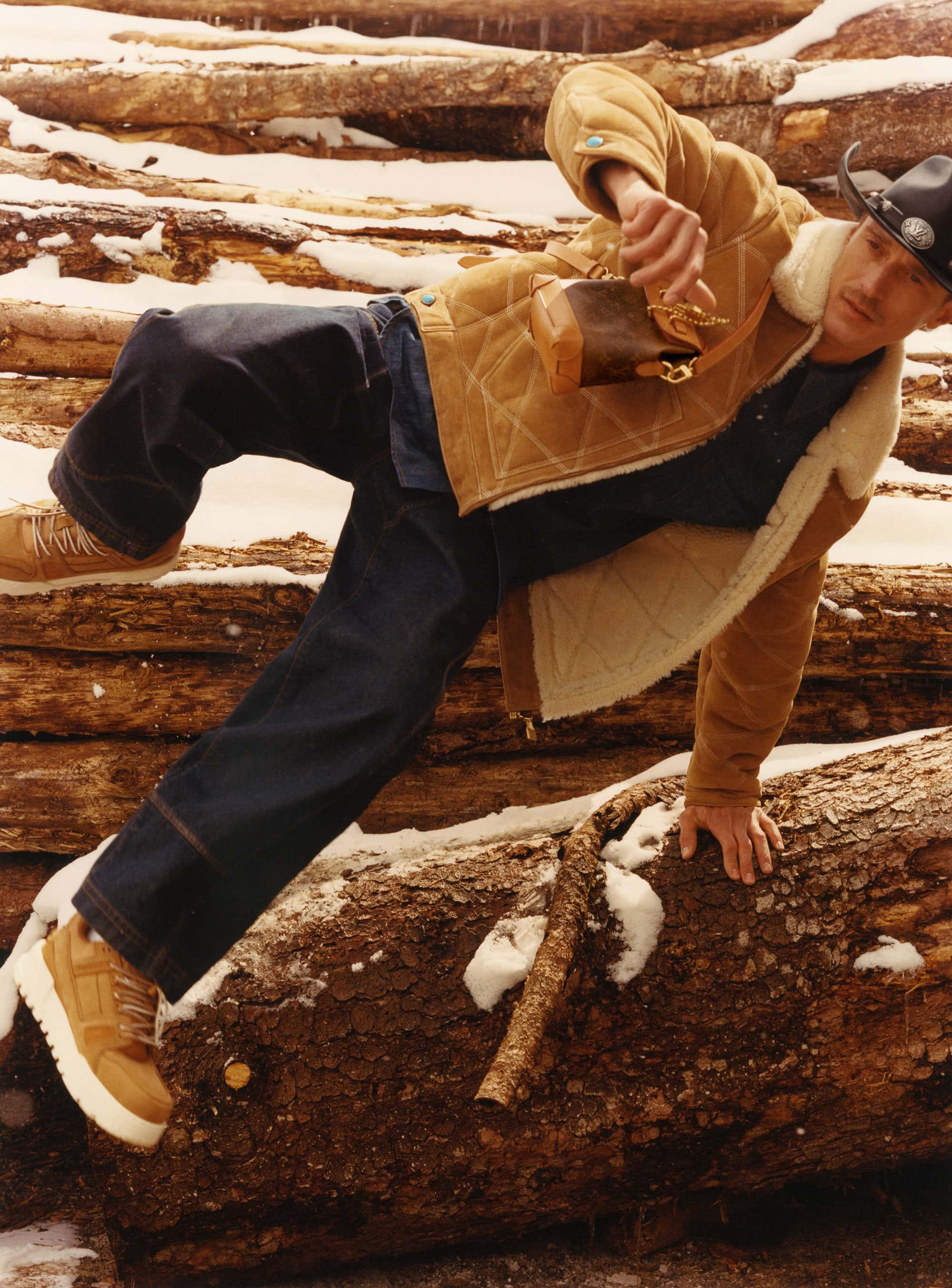 Man wearing a shearling-lined tan jacket, dark jeans, and cowboy hat climbing over snow-covered logs