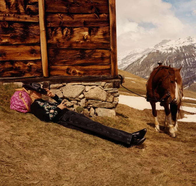 Woman in black outfit lies next to a log cabin and a horse in a rural landscape with mountains in the background on a sunny day.