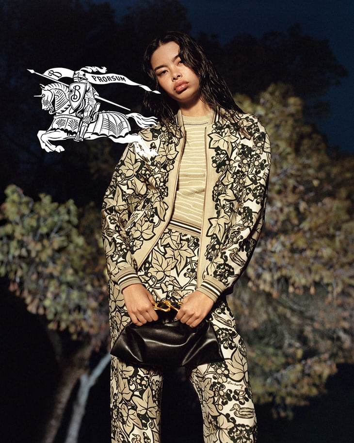 A stylish Asian woman poses outdoors in a beige patterned pantsuit, holding a black purse. A cartoon emblem floats in the background.
