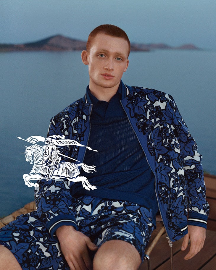 Male fashion model in floral jacket and shorts poses outdoors with blurred ocean backdrop. Fashion editorial featuring Burberry logo.