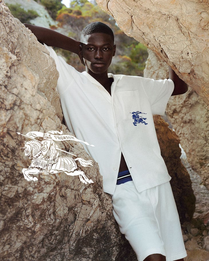 Young man in white shirt and shorts poses among rocks in sunlit outdoor fashion shot, wearing Burberry logo apparel in a natural setting.