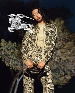 Model wearing patterned leaf-print jacket and trousers holding a black leather handbag against an outdoor nocturnal background