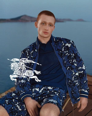 A red-haired male model wearing a blue patterned jacket and shorts seated outdoors against an ocean backdrop.