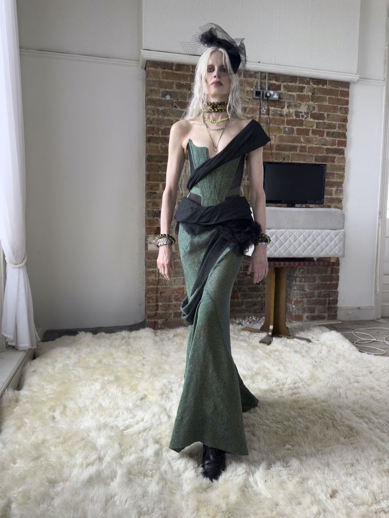 Elegant woman in green gown, indoor setting. She poses on a fluffy rug with a brick wall backdrop and vintage decor.