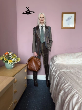 Model with platinum blonde hair wearing a striped suit and boots, holding a brown leather bag in a bedroom