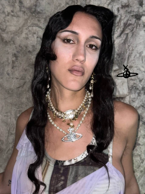 Close-up portrait of an adult woman with long dark hair wearing pearl necklaces and earrings against a textured wall background.