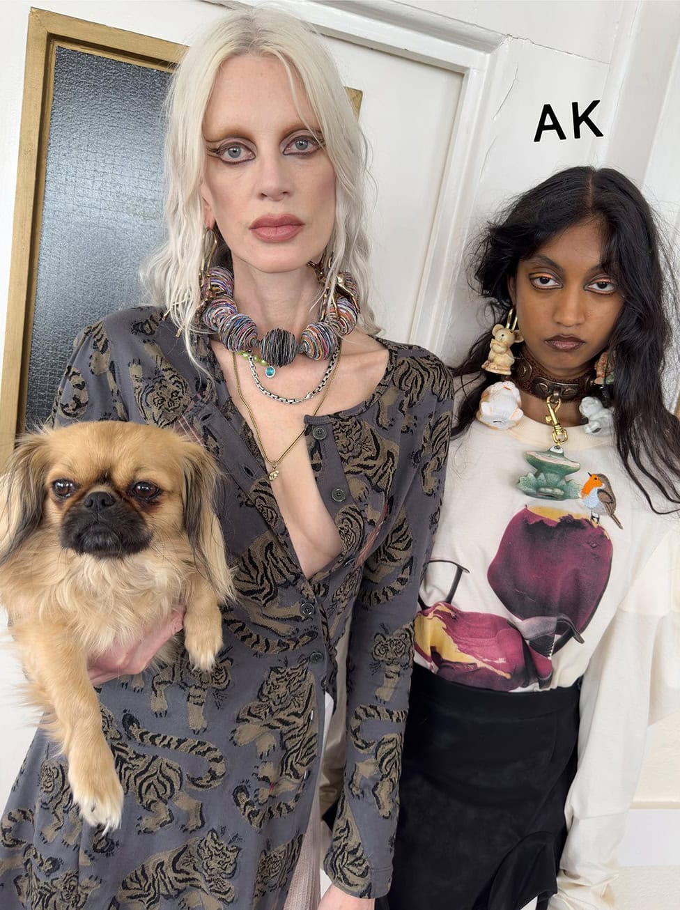 Two women pose indoors: one holds a tan-colored Pekingese dog; both wear statement jewelry and contemporary casual fashion with neutral backdrop.
