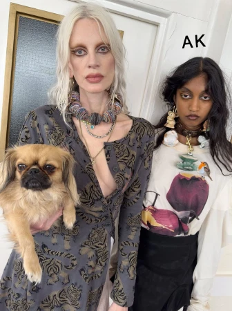 Two women pose indoors: one holds a tan-colored Pekingese dog; both wear statement jewelry and contemporary casual fashion with neutral backdrop.