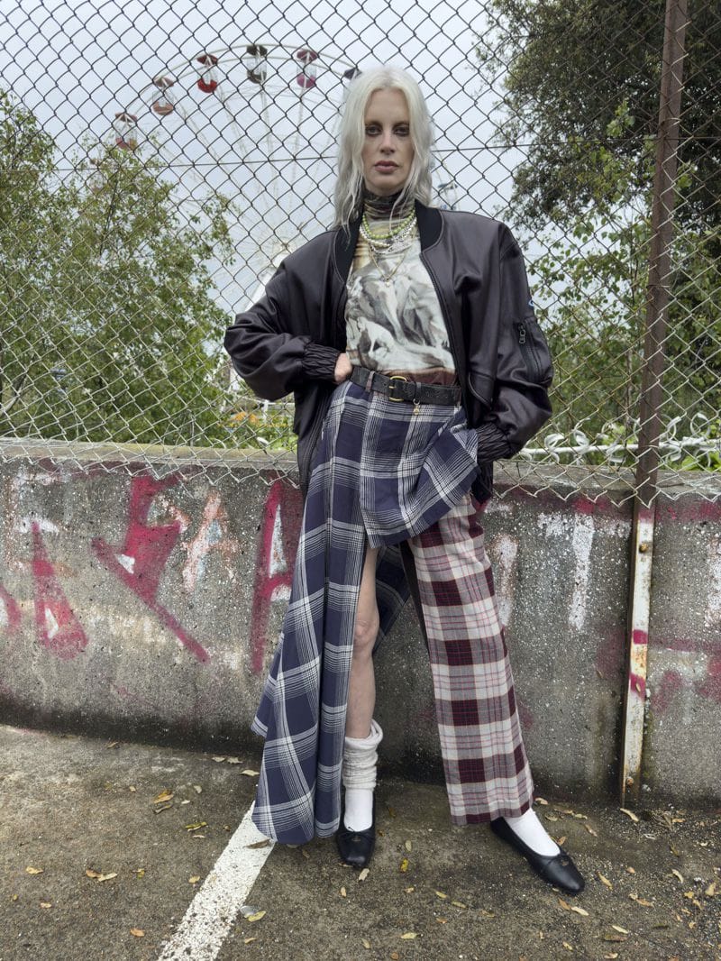 Fashionable young adult with platinum hair poses outdoors in a black jacket, plaid skirt, and black flats, near a chain-link fence.
