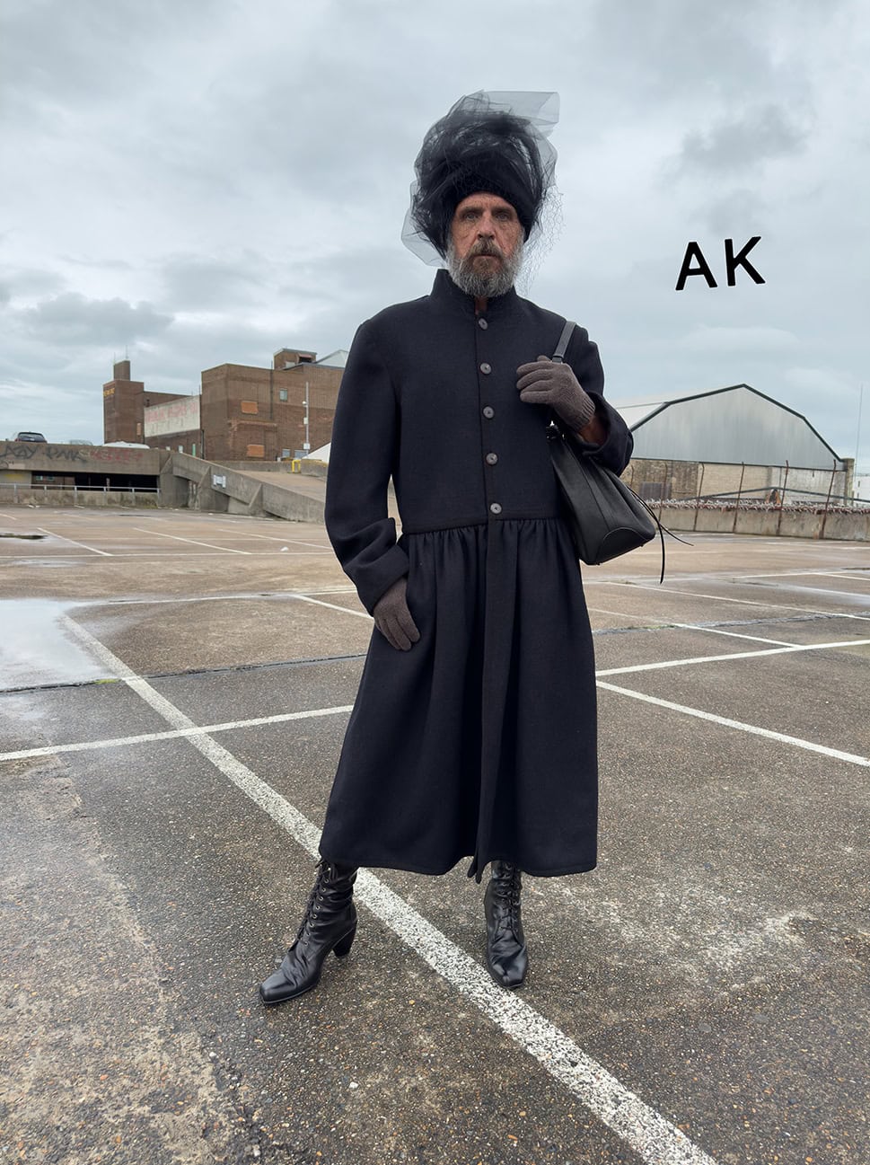 Person in dark coat with headdress and black purse stands on an outdoor parking lot with a building in the background on a cloudy day.