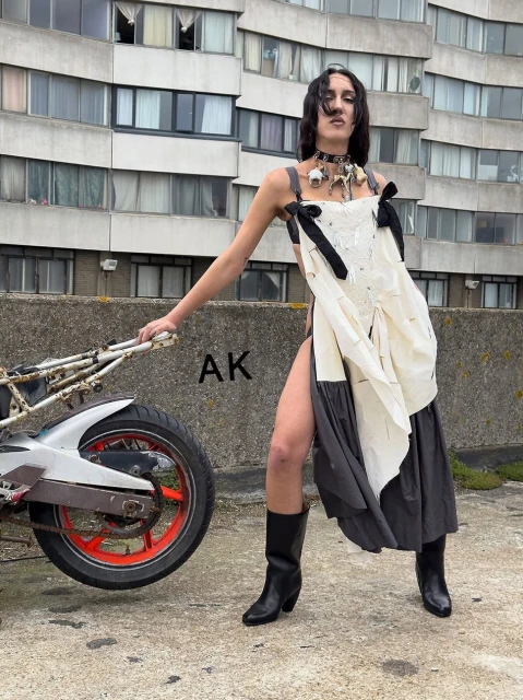 Adult female poses in avant-garde dress with motorcycle and urban building backdrop; edgy fashion photo showcases creative design.