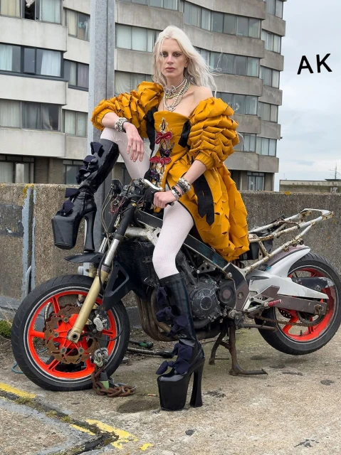 Woman in avant-garde fashion seated on a motorcycle against an urban building backdrop, wearing platform boots and jewelry.