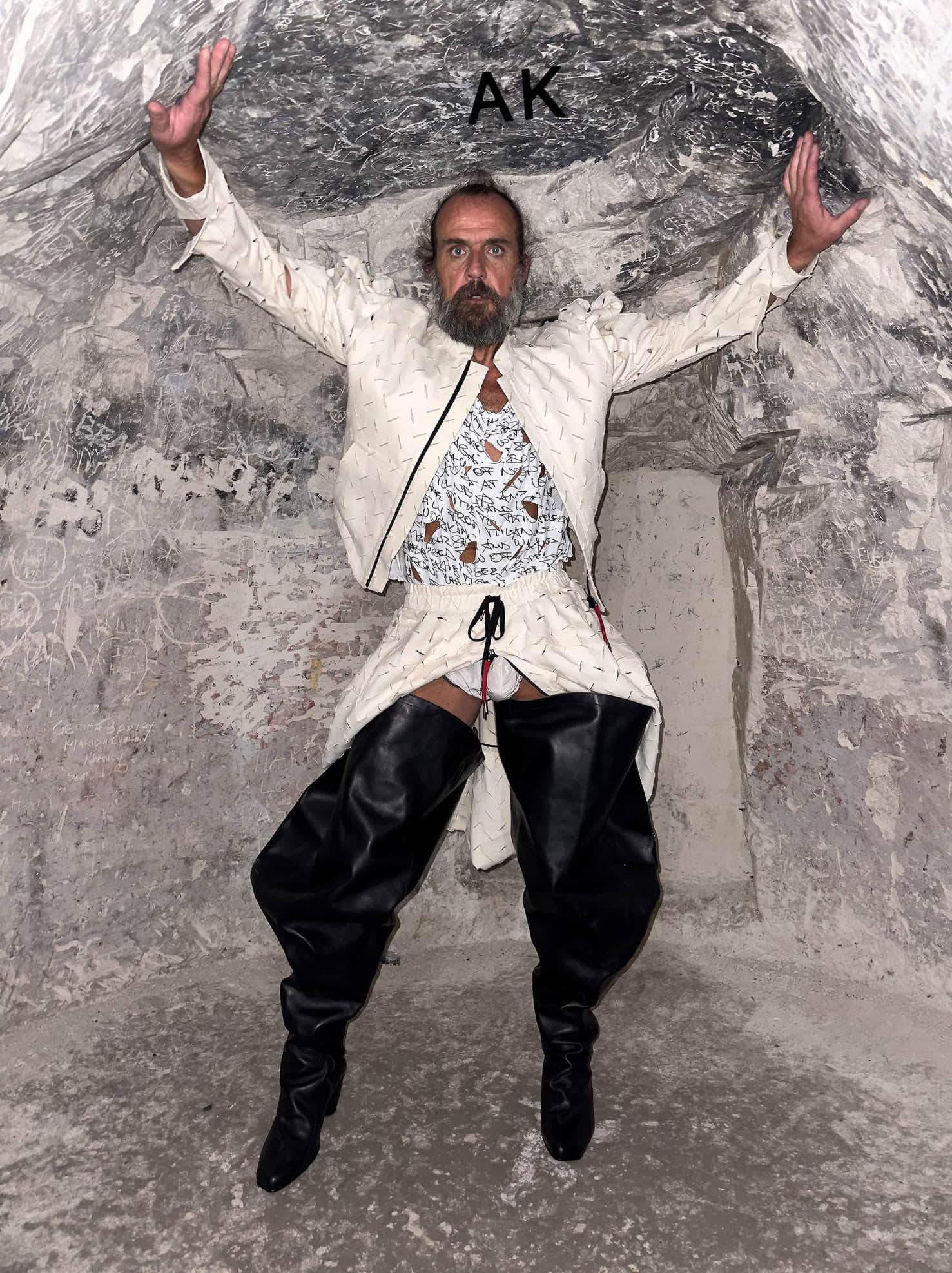 Adult male in a white suit and black boots stands with arms up in a stone cave with overhead light. High fashion with stone backdrop.