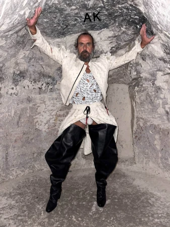 Adult male in a white suit and black boots stands with arms up in a stone cave with overhead light. High fashion with stone backdrop.