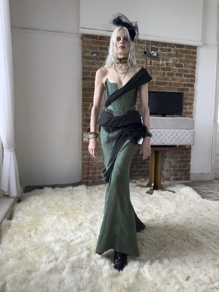 Model in green structured gown and black fascinator standing on white shaggy rug indoors