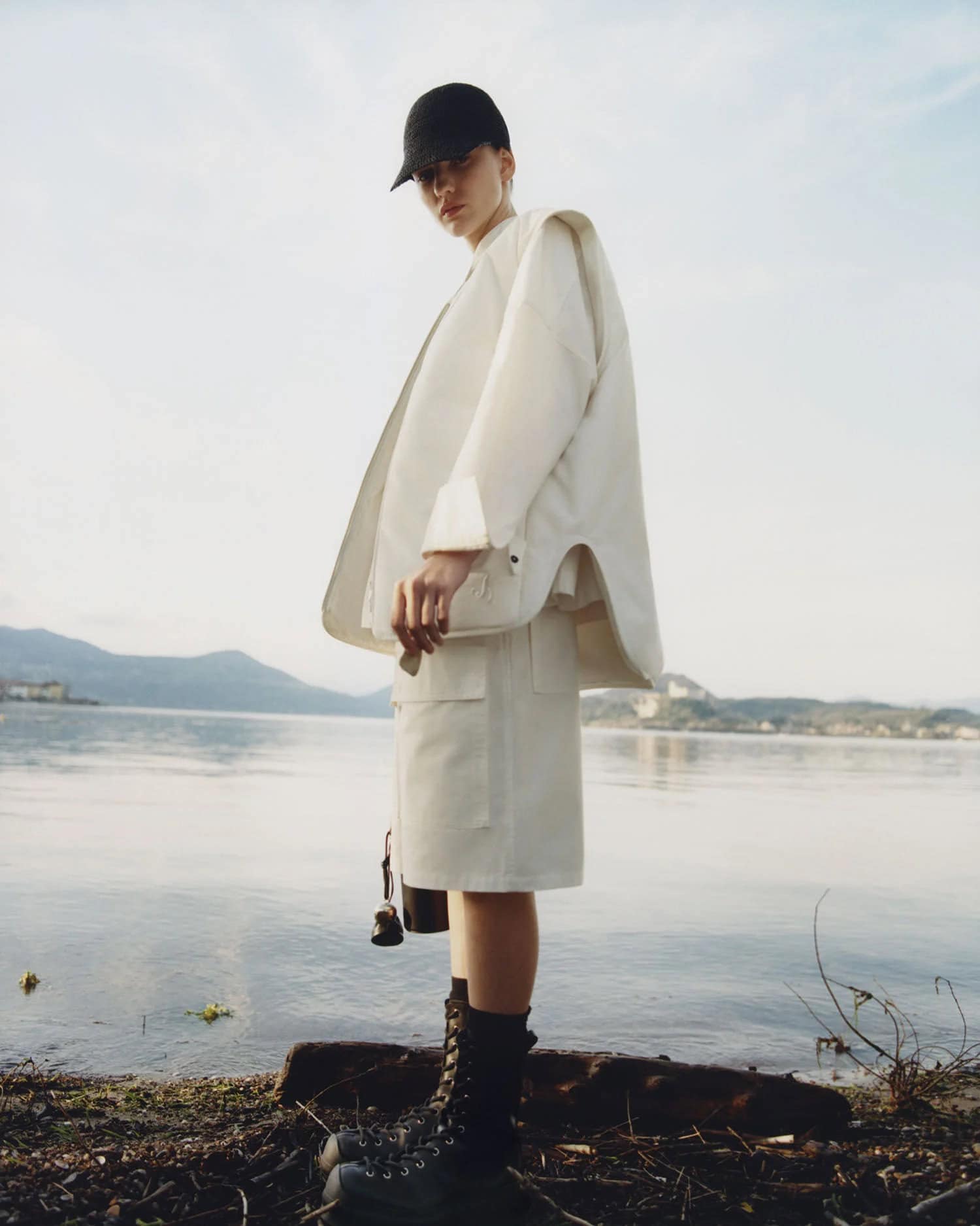 Fashionable woman in a baseball cap and white jacket and skirt standing on the bank of a lake with mountains in the distance.