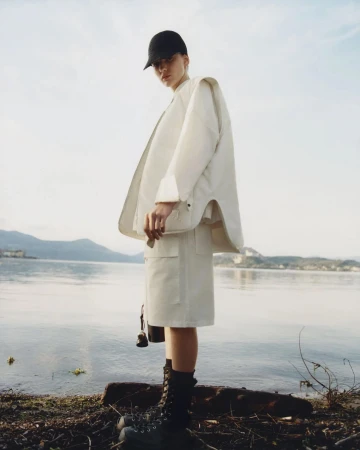 Fashionable woman in a baseball cap and white jacket and skirt standing on the bank of a lake with mountains in the distance.