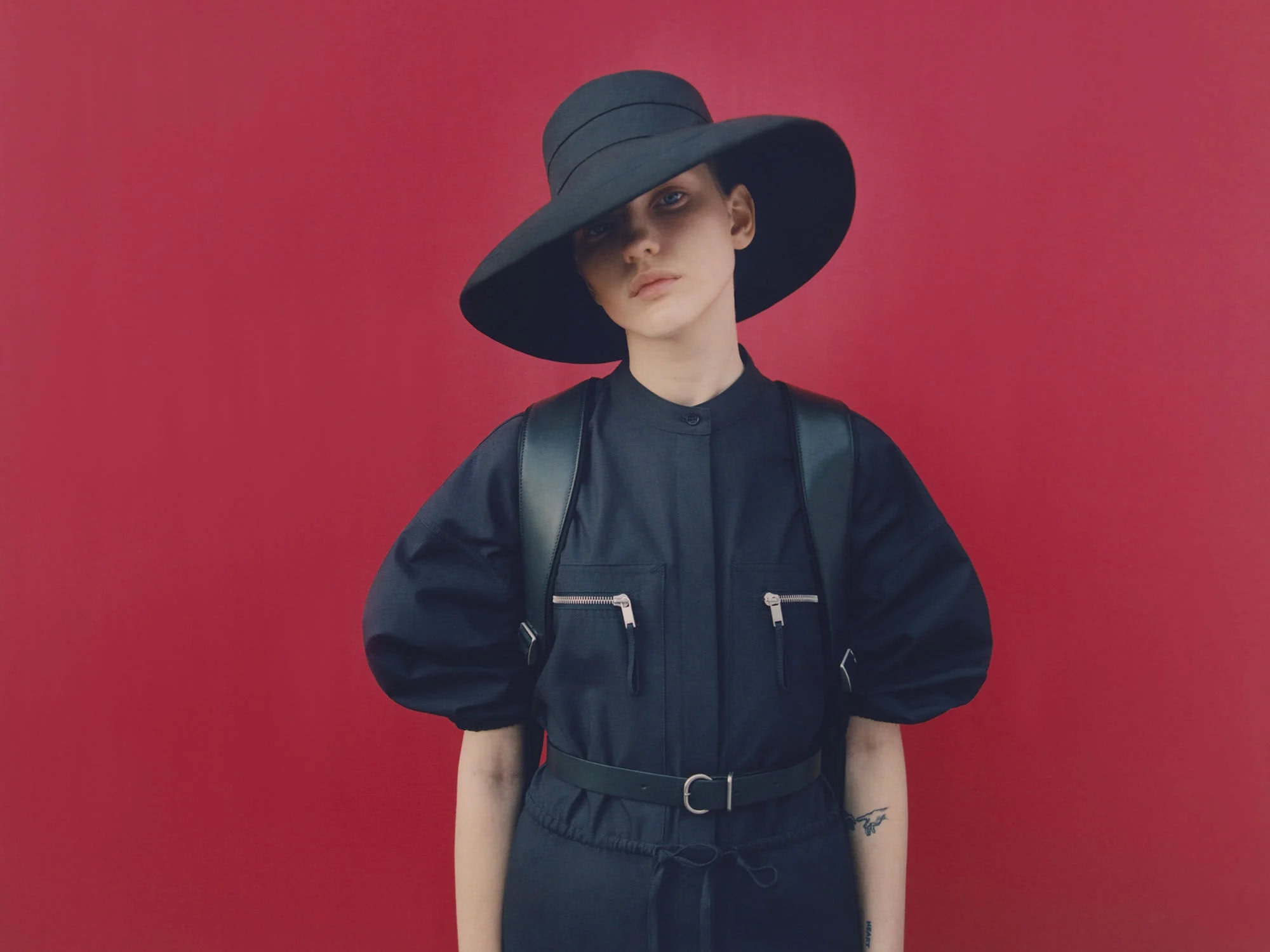 A young adult female poses in a dark jumpsuit, hat, and harness against a red backdrop in a studio fashion shot.