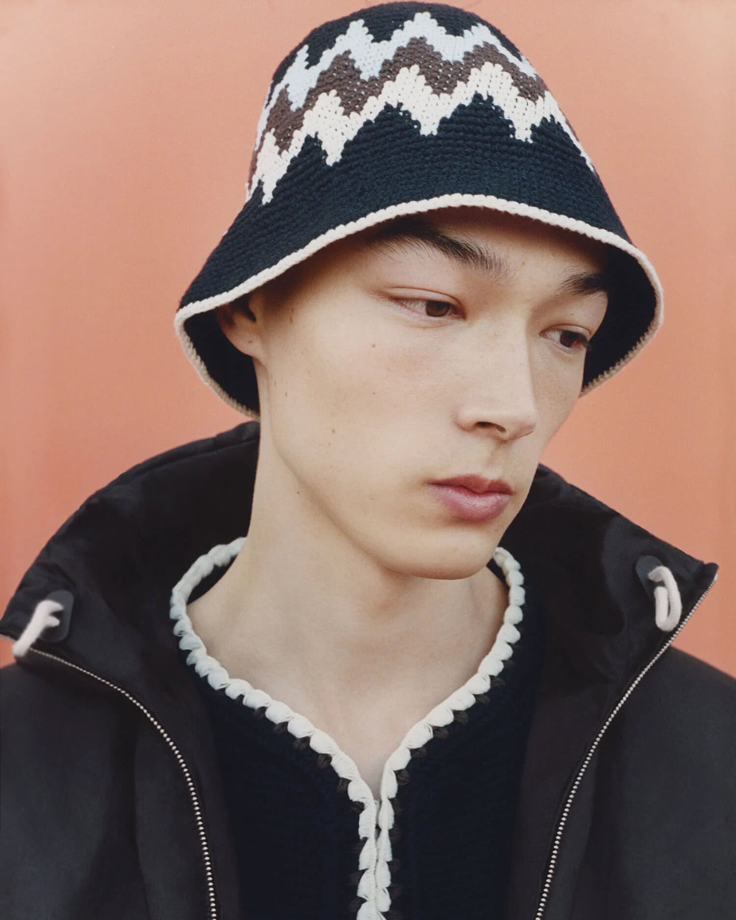 Portrait of a young adult male wearing a black jacket and a patterned bucket hat against an orange backdrop in a studio.