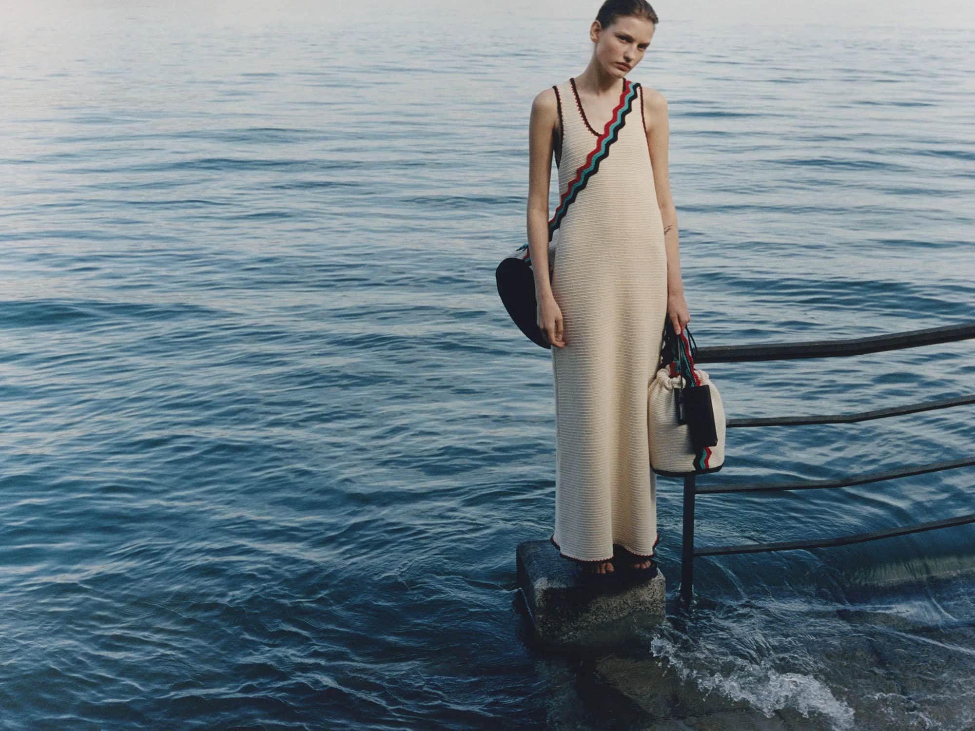 Adult woman in a long dress standing on a rock in water, holding a bag; railings and ocean visible, medium shot, outdoor setting.